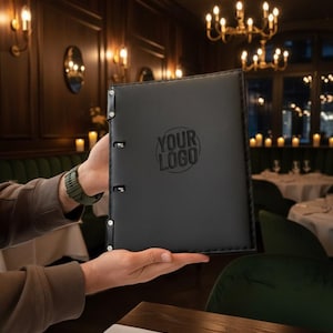 May include: Black leather menu cover with the text "YOUR LOGO" embossed on the front. The menu cover has a three-ring binder mechanism. The background shows a restaurant setting with tables and chairs.
