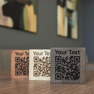 Puede incluir: Tres bloques de madera con códigos QR en tonos marrón, beige y gris. Cada bloque presenta un código QR único y el texto "Your Text" grabado. Los bloques se exhiben sobre una superficie de madera.