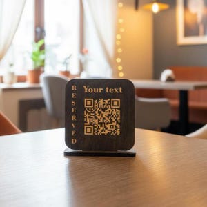 May include: A dark wooden table sign with the word "RESERVED" on the left side and "Your text" above a QR code. The sign is on a wooden table in a restaurant setting.
