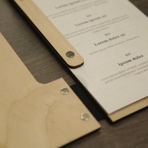 May include: A wooden menu holder with a black strap and metal fasteners. The menu is printed on white paper with the text "Lorem ipsum", "Lorem dolor sit", "Ipsum dolor", and prices in dollars.