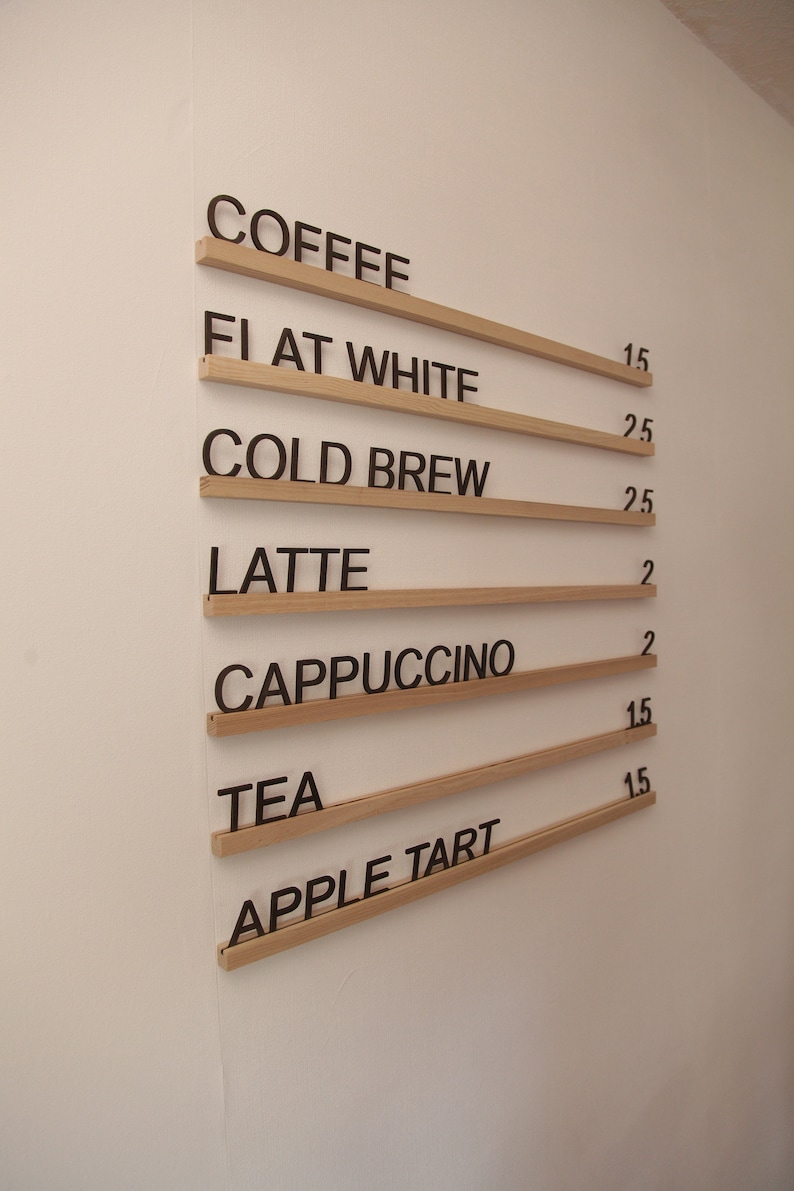 K&ouml;nnte beinhalten: Eine Holztafel mit wei&szlig;em Hintergrund listet Kaffee- und Teegetr&auml;nke mit Preisen auf. Die Speisekarte umfasst Kaffee, Flat White, Cold Brew, Latte, Cappuccino, Tee und Apfelkuchen.
