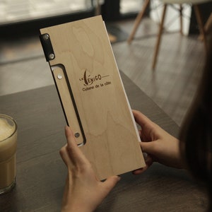May include: A wooden menu cover with a black leather strap and metal accents. The cover has the text "Le  ENIGO - Cuisine de la cite" engraved on it.