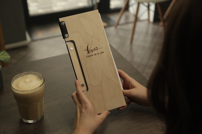 May include: A wooden menu cover with a black leather strap and metal accents. The cover has the text "Le  ENIGO - Cuisine de la cite" engraved on it.
