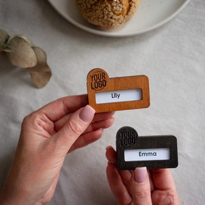 May include: Two wooden place card holders with a slot for a name card. The top holder is light brown with the text "YOUR LOGO" and the name "Lily". The bottom holder is dark brown with the text "YOUR LOGO" and the name "Emma".