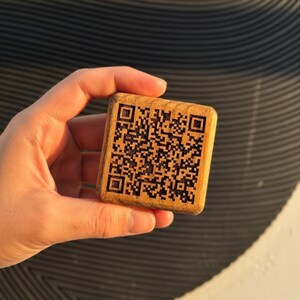 Puede incluir: Un bloque de madera cuadrado con un código QR grabado. El código es un patrón de cuadrados blancos y negros. El bloque se sostiene en una mano. La madera es de color marrón claro y tiene una veta natural.