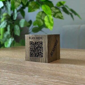 Puede incluir: Cubo de madera marrón con las palabras «SCAN MENU» y un código QR. La palabra «RESERVADO» está impresa en diagonal en un lado. El cubo está sobre una mesa de madera, con follaje verde en el fondo.