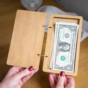 May include: A wooden journal with a built-in cash compartment. The compartment is open and contains several US dollar bills. The journal has a ring binder and is made of light brown wood.