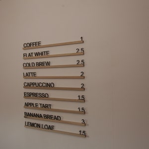 K&ouml;nnte beinhalten: Eine wei&szlig;e Wand mit einer Speisekarte, auf der Kaffee und Backwaren mit Preisen aufgef&uuml;hrt sind. Die Speisekarte ist in schwarzer Schrift auf Holzleisten geschrieben.