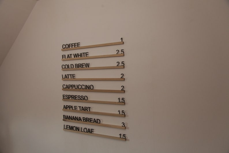 K&ouml;nnte beinhalten: Eine wei&szlig;e Wand mit einer Speisekarte, auf der Kaffee und Backwaren mit Preisen aufgef&uuml;hrt sind. Die Speisekarte ist in schwarzer Schrift auf Holzleisten geschrieben.