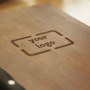 Puede incluir: Un diario de madera con una cubierta de cuero negro y tachuelas plateadas. El diario tiene las palabras "your logo" grabadas en la parte delantera.