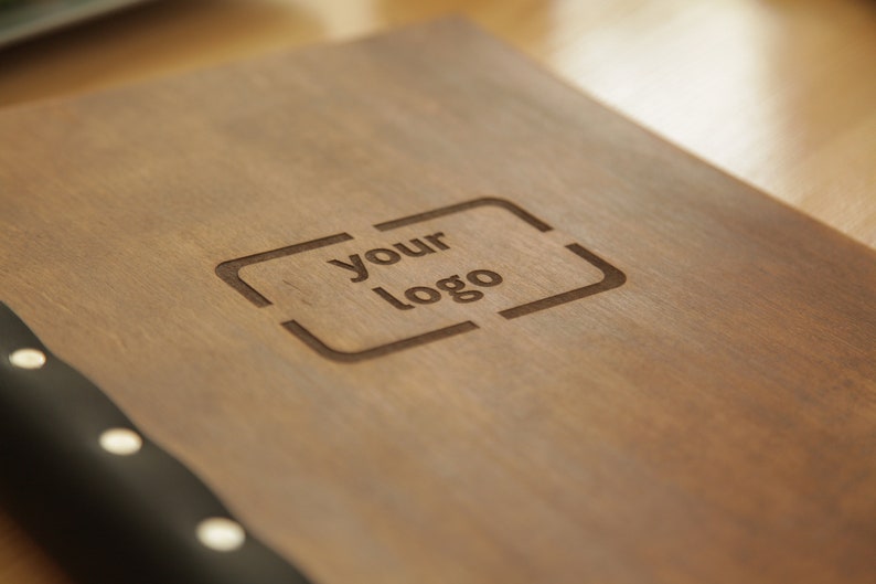 Puede incluir: Un diario de madera con una cubierta de cuero negro y tachuelas plateadas. El diario tiene las palabras "your logo" grabadas en la parte delantera.