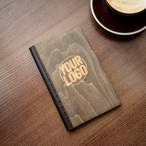 Puede incluir: Una cubierta de menú de madera con un borde de cuero negro y el texto "YOUR LOGO" en letras doradas.