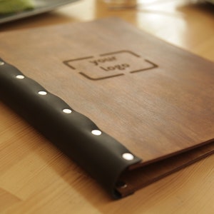 Puede incluir: Una cubierta de men&uacute; de madera marr&oacute;n con una columna vertebral de cuero negro y remaches plateados. La cubierta tiene un recorte rectangular con el texto "your logo" grabado en el interior.