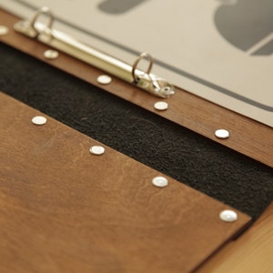 Puede incluir: Un archivador de madera marr&oacute;n con forro de tela negro y remaches de metal plateado. El archivador tiene un mecanismo de anillo de metal plateado.