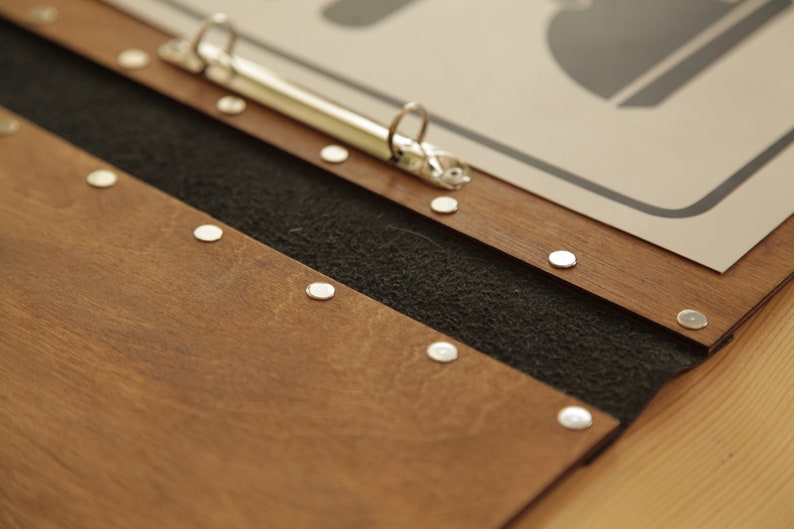 Puede incluir: Un archivador de madera marr&oacute;n con forro de tela negro y remaches de metal plateado. El archivador tiene un mecanismo de anillo de metal plateado.
