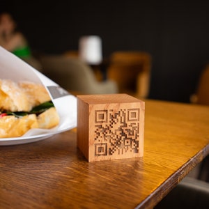 May include: A wooden block with a black and white QR code engraved on the front. The block is sitting on a wooden table.