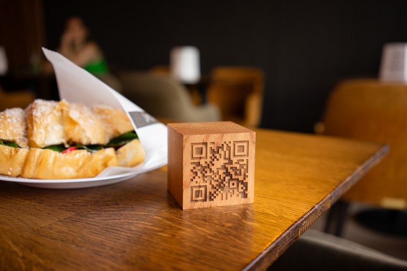 May include: A wooden block with a black and white QR code engraved on the front. The block is sitting on a wooden table.