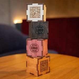 Könnte beinhalten: Ein Stapel aus vier Holzkisten mit unterschiedlichen Designs. Die oberste Kiste ist weiß mit einem schwarzen QR-Code, die zweite Kiste ist schwarz mit einem weißen Kreis, auf dem "YOUR LOGO HERE" steht, die dritte Kiste ist rosa mit einem weißen Kreis, auf dem "YOUR LOGO HERE" steht, und die unterste Kiste ist braun mit einem schwarzen QR-Code.