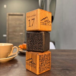 以下が含まれることがあります： 3つの木製テーブル番号ブロックが積み重ねられています。一番上のブロックは薄茶色で、数字の17と「Tirrenia」の文字が入っています。真ん中のブロックは濃い茶色でQRコードが入っています。一番下のブロックは薄茶色で、QRコードと「Tirrenia」の文字が入っています。
