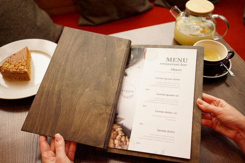 May include: A wooden menu with a white paper insert listing menu items for a restaurant-bar. The menu is open to reveal the text "MENU restaurant-bar Share" and a list of menu items with prices. The menu is on a wooden table with a plate of cake, a glass teapot, and a cup of tea.