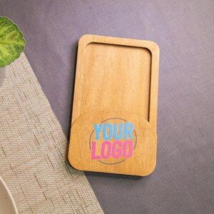 Op de afbeelding: Een houten telefoonstandaard met een uitsparing voor een telefoon en een cirkelvormig gebied met de tekst "YOUR LOGO" in roze en blauwe letters.