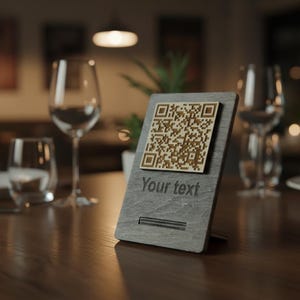 May include: A grey wooden table sign featuring a QR code and the text "Your text". The sign has a brown QR code and is displayed on a wooden table. The background includes wine glasses and a plant.