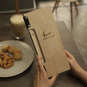 May include: A wooden menu with a black leather strap and metal rivets. The menu is engraved with the text "Le Yenigo - Cuisine de la cite".