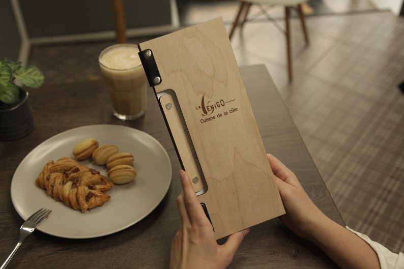 May include: A wooden menu with a black leather strap and metal rivets. The menu is engraved with the text "Le Yenigo - Cuisine de la cite".