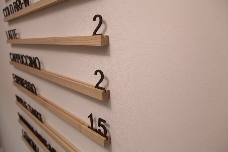 K&ouml;nnte beinhalten: Eine an der Wand montierte Speisekarte aus Holz mit einer Liste von Kaffeegetr&auml;nken und Zahlen, die die Menge jedes Getr&auml;nks angeben. Die aufgef&uuml;hrten Getr&auml;nke sind: Cold Brew, Latte, Cappuccino, Espresso, Double Shot, Americano und Lemon & Ginger.