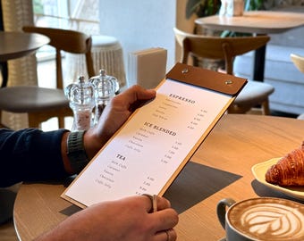 May include: A cafe menu with espresso, ice blended, and tea options. The menu is held by a person at a table with a cup of coffee and a croissant. The menu has a brown leather cover and is open to display the drink options.