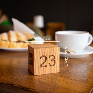 May include: A wooden table number cube with the number 23 engraved on it. The cube is 6 cm / 2.4 inches wide. The cube is on a wooden table with a cup of coffee and a croissant in the background.