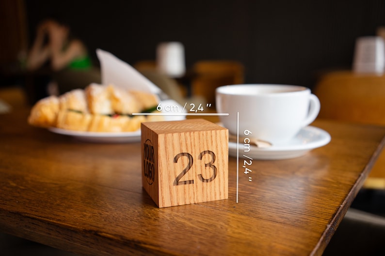 May include: A wooden table number cube with the number 23 engraved on it. The cube is 6 cm / 2.4 inches wide. The cube is on a wooden table with a cup of coffee and a croissant in the background.