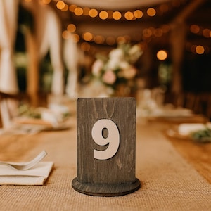 May include: A wooden table number nine, with a light beige number on a dark brown rectangular base. The table number is on a table with a burlap runner, with a blurred background of lights and floral arrangements.