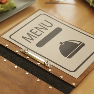Puede incluir: Una cubierta de men&uacute; de madera con una columna vertebral negra y un archivador de anillas de metal. La cubierta tiene un inserto de papel blanco con la palabra "MENU" y un icono en blanco y negro de una campana.