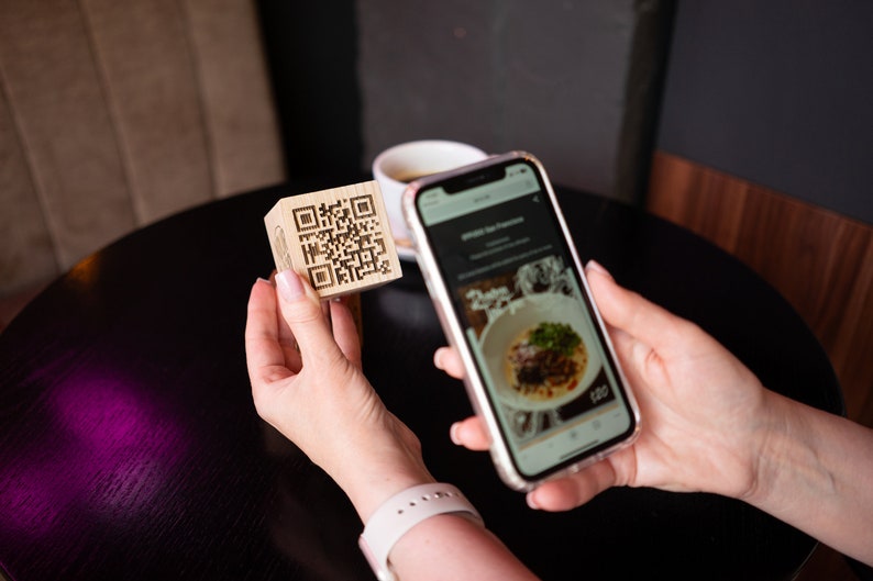 May include: A person uses a smartphone to scan a QR code on a wooden block. The phone screen shows a menu with a photo of a dish and the price $12.00.