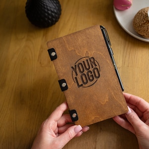May include: A brown wooden notebook with a black pen and the text "YOUR LOGO" engraved on the cover.