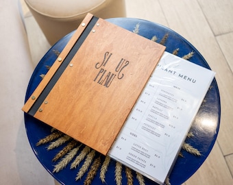 Puede incluir: Una cubierta de menú de madera con una correa de cuero negro y el texto "Si tu peux" en la parte delantera. El menú está abierto para revelar un menú de papel blanco con el texto "Restaurant Menu" y una lista de artículos de comida.