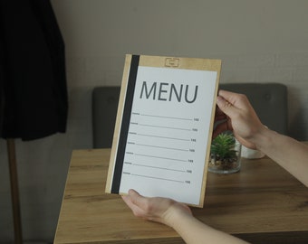 Könnte beinhalten: Ein Holz-Menüklemmbrett mit einem schwarzen Gummiband und dem Wort "MENU" in Schwarz auf einem weißen Blatt Papier. Das Klemmbrett hat einen leeren Platz zum Schreiben von Preisen und Beschreibungen.