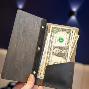 以下が含まれることがあります： 黒い革のフラップが付いた木製の財布で、開くとアメリカドル紙幣がいくつか見えます。財布は濃い木目調で、縁に金属製の留め具がいくつか付いています。