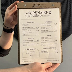 May include: A menu from "Aldenaire & Partners" restaurant, held by hands. The menu features a brown leatherette cover with metal rivets. The menu lists main dishes, appetizers, desserts, drinks, and additional toppings.