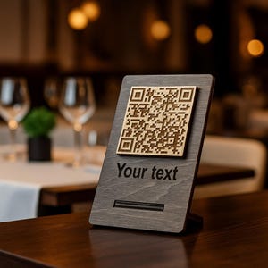 Peut inclure: Panneau en bois gris foncé avec un code QR en bois clair et le texte "Your text" en dessous. Le panneau est incliné et conçu pour être posé sur une table, idéal pour un restaurant.