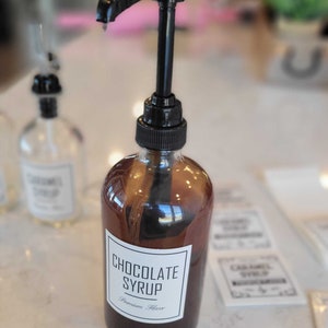 May include: A glass bottle with a black pump dispenser filled with dark brown chocolate syrup. The label reads "Chocolate Syrup" and "Premium Flavor".