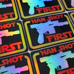 May include: A close-up of several holographic stickers with the text "HAN SHOT FIRST" in a yellow and red font. The stickers feature a silhouette of a blaster pistol.