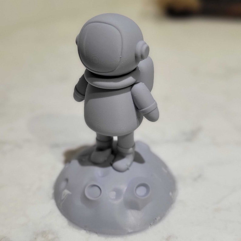 Pode incluir: Uma figura de astronauta cinza impressa em 3D em p&eacute; em uma base lunar cinza impressa em 3D.