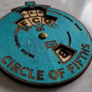 Puede incluir: Una herramienta de teor&iacute;a musical de madera, un c&iacute;rculo de quintas, con un acabado te&ntilde;ido de azul. El c&iacute;rculo tiene un dial giratorio con las notas C, D, E, F, G, A y B. El dial est&aacute; configurado en la nota B.
