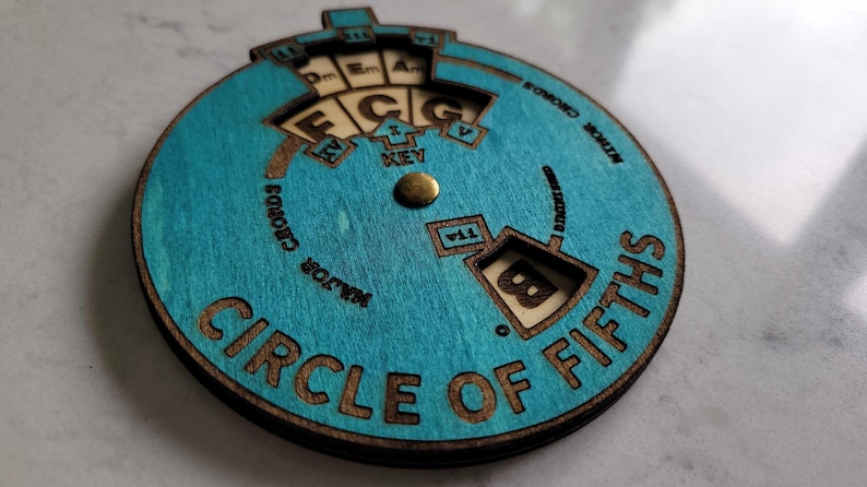 Puede incluir: Una herramienta de teor&iacute;a musical de madera, un c&iacute;rculo de quintas, con un acabado te&ntilde;ido de azul. El c&iacute;rculo tiene un dial giratorio con las notas C, D, E, F, G, A y B. El dial est&aacute; configurado en la nota B.