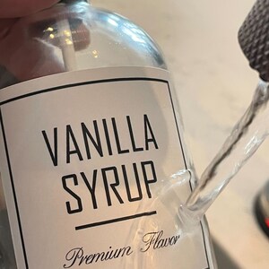 May include: A clear glass bottle with a white label that says "Vanilla Syrup Premium Flavor". The bottle is being poured into a clear glass container.