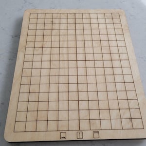 May include: A rectangular wooden board with a grid pattern, likely for a game. The board is light brown with dark brown grid lines and three small symbols at the bottom. The board is on a light-colored surface.
