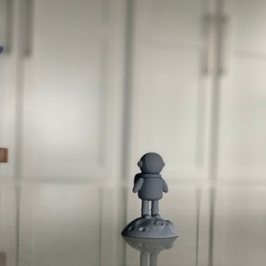 Pode incluir: Uma figura de astronauta cinza em p&eacute; sobre uma base cinza em forma de lua. O astronauta est&aacute; usando um capacete e tem um design simples.