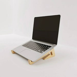 Può includere: Un computer portatile è posizionato su un supporto per laptop in legno. Il supporto è in legno marrone chiaro e ha una forma triangolare. Il laptop è argentato e ha uno schermo nero.